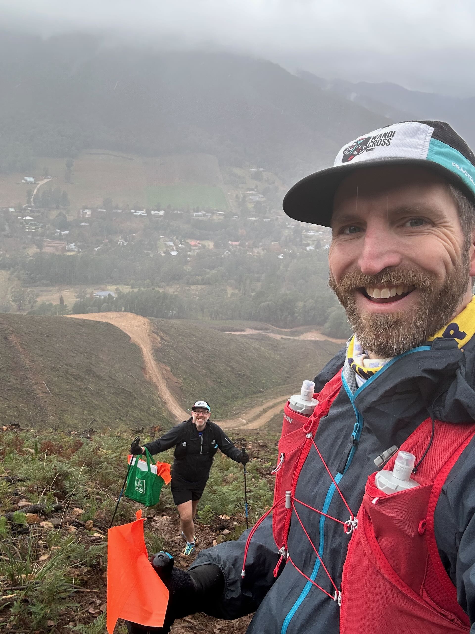 A selfie of Ross and his running mate Darren. They're climbing a steep hill in the clouds, with a bag of orange race flags they have collected whilst volunteering at a trail running race.